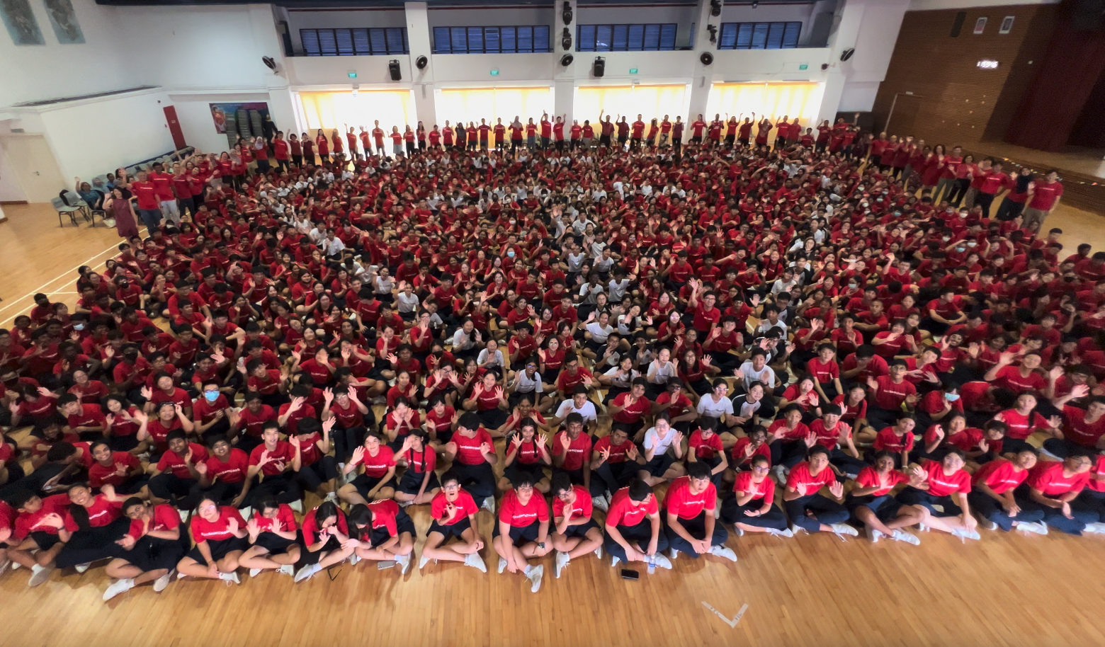 Large group of hundreds of students in red shirts gathered in a school gymnasium, waving at the camera.
